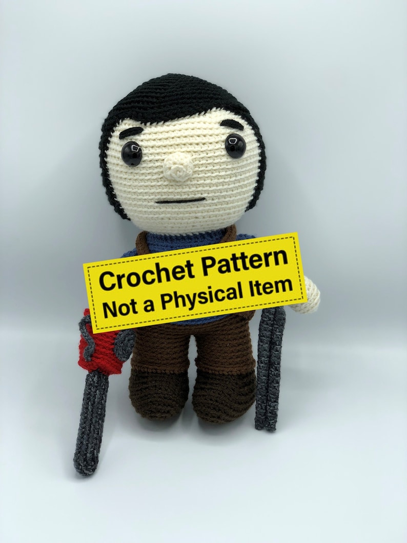 K&ouml;nnte beinhalten: Geh&auml;kelte Puppe mit schwarzen Haaren, cremefarbenem Gesicht und brauner Hose. Die Puppe h&auml;lt eine graue Kettens&auml;ge in der einen und eine graue Waffe in der anderen Hand. Ein gelbes Schild zeigt "Crochet Pattern Not a Physical Item."
