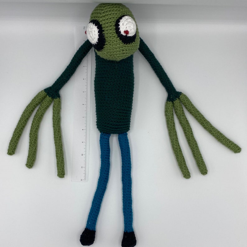 May include: Green crocheted plush toy with large eyes, green arms, and blue legs. The toy is approximately 30 cm tall.