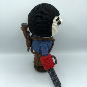 K&ouml;nnte beinhalten: Geh&auml;kelte Puppe eines Charakters mit schwarzer M&uuml;tze, blauem Hemd und braunem Geschirr. Die Puppe hat einen Kettens&auml;genarm mit rotem Griff und eine Schrotflinte. Einzigartiges Design, handgefertigt.
