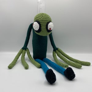 May include: Green crocheted plush toy with large white eyes and red pupils. The toy has long green arms and legs, with blue trousers and black shoes.