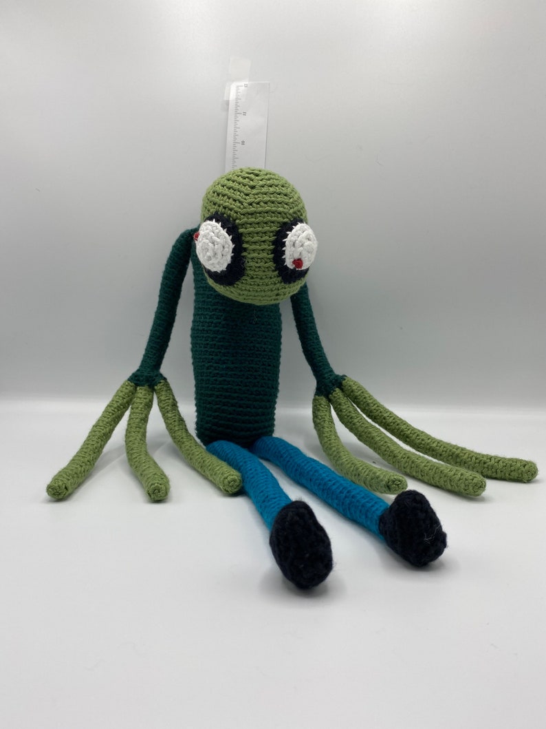 May include: Green crocheted plush toy with large white eyes and red pupils. The toy has long green arms and legs, with blue trousers and black shoes.