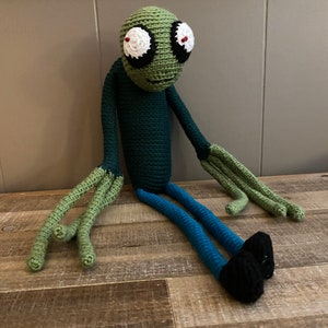May include: Green crocheted plush toy with large eyes, a green body, blue trousers, and black shoes. The toy is sitting on a wooden surface.
