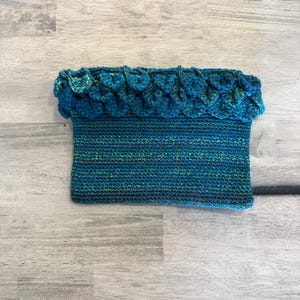 May include: A handmade, teal-colored crochet cowl with a scalloped edge. The cowl is a rectangle shape with a textured stitch pattern. The top edge features a row of teal-colored, scale-like embellishments. The yarn has flecks of green and yellow.