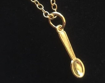 May include: A gold-colored necklace featuring a delicate chain and a small, singular spoon-shaped pendant. The pendant is attached to the chain by a circular jump ring, and the entire piece has a polished, metallic finish with a bright yellow hue.