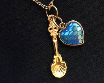 May include: A gold-toned metal necklace featuring a delicate chain with two charms attached to a jump ring. One charm is a small gold spoon with a shell-shaped bowl, and the other is a heart-shaped pendant with a blue iridescent scale pattern.