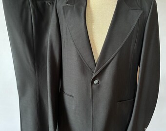 Könnte beinhalten: Ein schwarzer Anzug, bestehend aus Jacke und Hose, auf einer Schaufensterpuppe präsentiert. Die Jacke hat ein fallendes Revers, einen Ein-Knopf-Verschluss und zwei Vordertaschen. Die Hose hat ein gerades Bein. Der Anzug ist aus dunklem Stoff gefertigt.