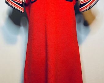 Könnte beinhalten: Ein rotes Vintage-Kleid mit V-Ausschnitt und kurzen Ärmeln. Das Kleid hat marine-, weiß- und rot gestreifte Akzente an Kragen, Ärmelbündchen und Saum. Zwei Taschen mit marinefarbenem Besatz befinden sich auf der Brust.