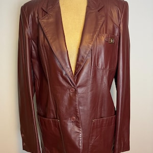 May include: A burgundy leather blazer with a notched lapel, two front pockets, and a single-breasted button closure. The blazer is displayed on a mannequin and features a small decorative detail on the pocket.