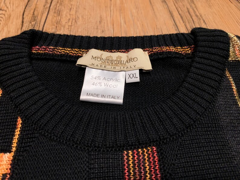Peut inclure: Pull-over noir &agrave; rayures orange, jaune et rouge. L'&eacute;tiquette indique "Montechiaro Made in Italy" et "54% Acrylique 46% Laine" avec une &eacute;tiquette de taille indiquant "XXL".