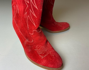Könnte beinhalten: Ein Paar rote Wildleder-Cowboystiefel. Die Stiefel haben weiße Nähte in einem dekorativen Muster. Die Sohlen sind braun. Die Stiefel sind zum Betrachter hin abgewinkelt.