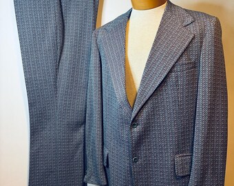 Könnte beinhalten: Ein zweiteiliger Anzug bestehend aus einem Blazer und einer passenden Hose mit dunkelblau-weißem Fischgrätenmuster. Der Blazer hat ein steigendes Revers, zwei Knöpfe auf der Vorderseite und zwei Pattentaschen. Die Hose hängt an einem Kleiderbügel.