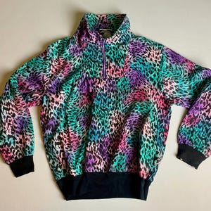 May include: A colorful pullover with a vibrant leopard print in shades of pink, purple, teal, and black. The long-sleeved top features a quarter-zip collar, black cuffs, and a black hemline. A retro-style garment.