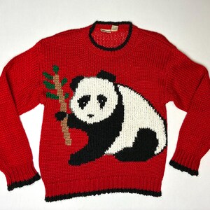 May include: A red knit sweater featuring a large panda bear design. The panda is black and white, holding a bamboo stalk with green leaves. The sweater has a crew neck and black trim on the collar, cuffs, and hem.