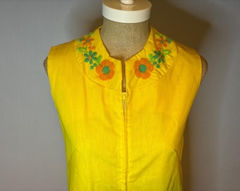 Könnte beinhalten: Ein ärmelloses, leuchtend gelbes Kleid mit rundem Kragen. Der Kragen ist mit gestickten Blumen in Orange, Grün und Gelb verziert. Ein goldener Reißverschluss verläuft über die Vorderseite des Kleides. Das Kleid wird auf einer Schaufensterpuppe präsentiert.