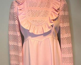 Könnte beinhalten: Ein hellrosa Vintage-Kleid mit hohem Rüschenkragen und langen Ärmeln. Das Oberteil hat ein strukturiertes, offenes Webmuster mit Rüschenbesatz. Die Ärmel haben ein ähnliches offenes Webmuster. Das Kleid hat eine taillierte Taille und einen fließenden Rock.