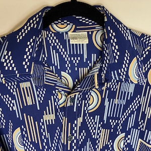 May include: A vintage button-down shirt in navy blue with a geometric pattern of white, light blue, and yellow lines and shapes. The shirt has a classic collar and a label with text.