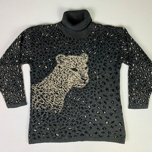 May include: A gray turtleneck sweater with a white leopard print design. The sweater features a large leopard head in the center.