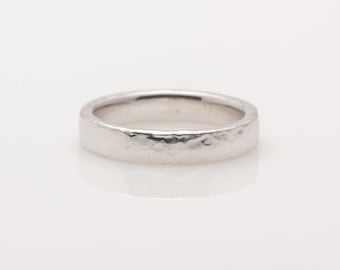 May include: A silver ring with a hammered texture. The band is a simple, flat design with a slightly irregular surface. The ring is set against a white background, highlighting its metallic sheen.