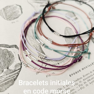 Peut inclure: Plusieurs bracelets de code Morse initial en noir, marron, violet, rose et turquoise. Chaque bracelet est orné de petites perles argentées et d'un détail cylindrique argenté. Ils sont présentés sur une page de livre vintage.