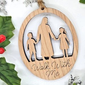 May include: Wooden Christmas ornament with a cutout design of a figure holding hands with two children. The text "Walk With Me" is engraved on the ornament. The ornament is round with a natural wood finish and a twine loop for hanging.