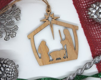 Könnte beinhalten: Ein hölzerner Weihnachtsbaumschmuck, der die Weihnachtsgeschichte darstellt. Der Anhänger ist in Form eines Stalls mit einem Stern darüber gestaltet. Die Figuren von Maria, Josef und dem Jesuskind sind aus dem Holz ausgeschnitten.