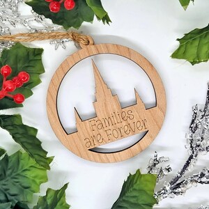 May include: A round wooden Christmas ornament with a cut-out design of a building silhouette and the words "Families are Forever." The ornament is surrounded by green leaves, red berries, and silver glitter.