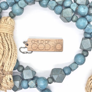 May include: A wooden keychain with the text "CHILD OF GOD" and a sun graphic. The keychain is attached to a silver chain and is surrounded by blue and teal beads.