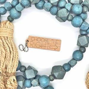 May include: A decorative wooden bead garland in shades of blue and gray, with various geometric shapes and round beads. A small wooden tag with the text "CHOOSE THE RIGHT" and an arrow is attached. A natural rope tassel is also visible.