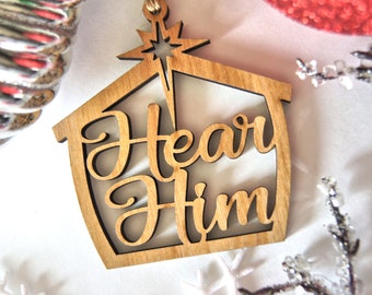 Könnte beinhalten: Weihnachtsbaumschmuck aus Holz in Form eines Hauses mit einem Stern oben. Der Anhänger sagt "Hear Him" in einer dekorativen Schrift.