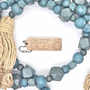 May include: A wooden keychain with the text "CHILD OF GOD" and a sun graphic. The keychain is attached to a silver chain and is surrounded by blue and teal wooden beads.