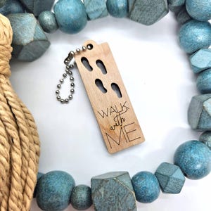 Puede incluir: Un llavero de madera con la inscripción "WALK with ME" grabada, con huellas recortadas. El llavero está sujeto a una cadena de bolas plateada. El fondo incluye cuentas de madera azules y turquesas y una cuerda.