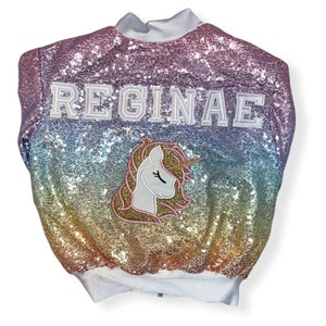 May include: A rainbow sequined jacket with a white zipper and the name "REGINAE" in white letters. A white unicorn with a gold horn and pink mane is embroidered on the front.