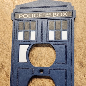 May include: A navy blue wall plate cover shaped like a police call box, with the words "POLICE PUBLIC CALL BOX" across the top. The cover has two circular openings for outlets and decorative details.