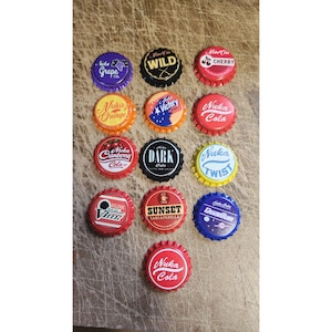 May include: A collection of thirteen circular metal bottle caps featuring various colourful graphic designs and text including Nuka Grape, Wild, Cherry, Orange, Victory, Nuka Cola, Cranberry, Dark, Twist, Vim, Sunset Sarsaparilla, Quantum, and Nuka Cola.