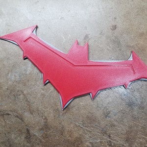 May include: A red bat-shaped emblem with a textured surface. The emblem has a gray outline and a central design element. The design is reminiscent of a superhero symbol.