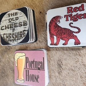 May include: Three stacks of square coasters. One stack features a circular design with the text THE OLD CHEESE OF CHESHIRE. Another stack shows a red tiger illustration with the text Red Tiger. The third stack displays a beer glass and the text Portugal House.