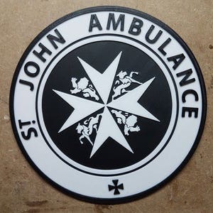 Könnte beinhalten: Ein kreisförmiges Emblem mit dem Text "ST JOHN AMBULANCE" in weißen Buchstaben. Das Emblem zeigt ein schwarz-weißes Malteserkreuz-Design mit Löwen und einem kleineren Kreuz unten. Der Rand ist schwarz.