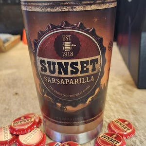May include: A stainless steel tumbler with a brown and red Sunset Sarsaparilla design. The tumbler has a clear lid and is surrounded by red bottle caps with the Sunset Sarsaparilla logo.