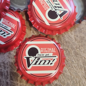 May include: Red bottle caps with the word "Vim!" in a stylized font. The caps have a red and white striped design with the word "Original" printed on them. The caps are scattered on a wooden surface.