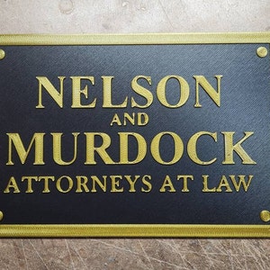 May include: A rectangular sign with the text "NELSON AND MURDOCK ATTORNEYS AT LAW" in gold lettering. The sign has a black background with a gold border and gold rivets in each corner. The sign is likely for a law firm.