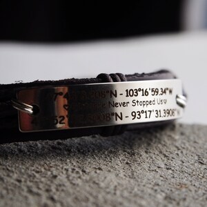 May include: A brown leather bracelet with a silver metal plate engraved with coordinates and the text "Never Stopped Us".