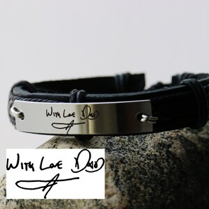 以下が含まれることがあります： 「With Love Dad」と手書きの署名で刻まれた銀色の金属プレートが付いた黒い革のブレスレット