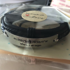 May include: Black leather bracelet with a silver plate engraved with the coordinates "40.5551&deg;N 75.9825&deg;W" and the text "Where it all began". The bracelet has a small heart charm with the words "handmade with love" on it.