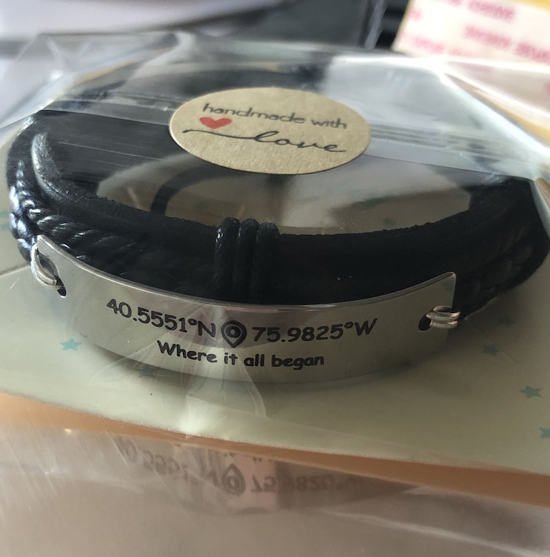 May include: Black leather bracelet with a silver plate engraved with the coordinates "40.5551&deg;N 75.9825&deg;W" and the text "Where it all began". The bracelet has a small heart charm with the words "handmade with love" on it.