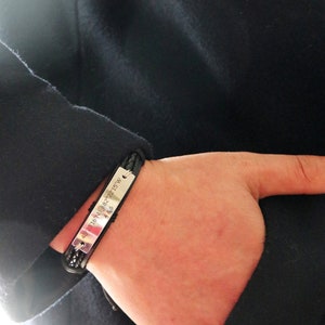 May include: A black leather bracelet with a silver plate engraved with the coordinates "40.2316&deg; N 82.2115&deg; W".