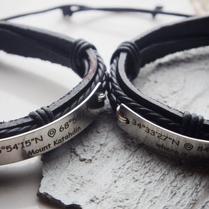 May include: Two black leather bracelets with silver metal plates engraved with coordinates and text. The top bracelet reads "45°54′15″N 68°55′17 Mount Katahdin". The bottom bracelet reads "34°33′27″N 84°15'08" where it all".