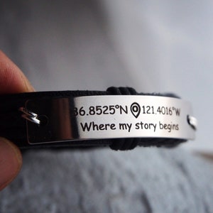 May include: Black leather bracelet with a silver metal plate. The plate is engraved with coordinates and the phrase "Where my story begins". The bracelet is a stylish accessory.