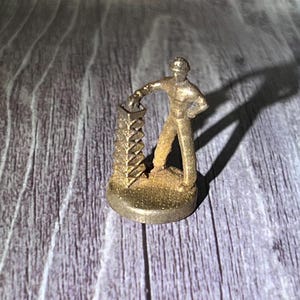 May include: A small, gold-colored figurine of a person in work clothes, standing next to a tall, detailed structure. The figure has one hand on the structure and the other on their hip. The base is round.