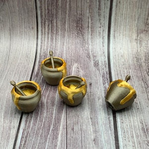 May include: Four small, gray ceramic honey pots with gold accents. Each pot has a small wooden dipper inside. The pots are arranged on a wooden surface.