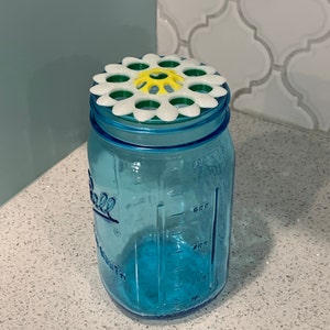 May include: A blue glass jar with a white and green daisy-shaped lid. The lid has a yellow centre with a green design.